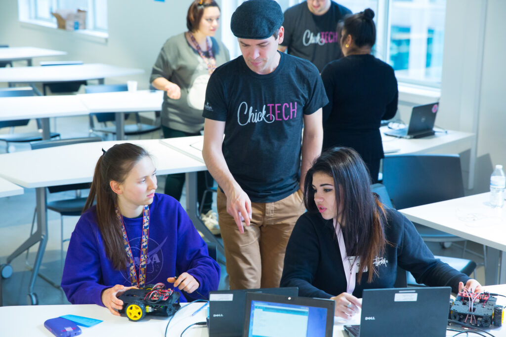 A male adult volunteer instructs two high school girls during a ChickTech robotics workshop.